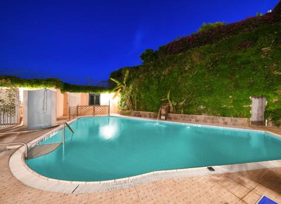 Piscina illuminata di notte, circondata da vegetazione rigogliosa e un cielo blu intenso.