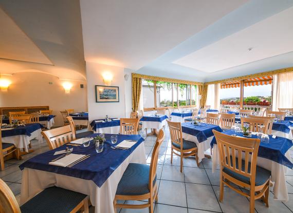 Ristorante luminoso, tavoli eleganti con tovaglie blu, atmosfera accogliente e vista panoramica.