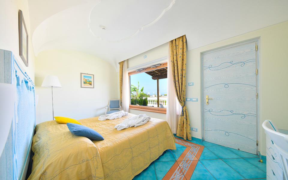 Camera luminosa con letto matrimoniale, vista mare e decorazioni blu e gialle.