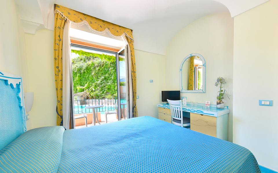 Camera luminosa con letto blu, vista su giardino e piscina, atmosfera rilassante.