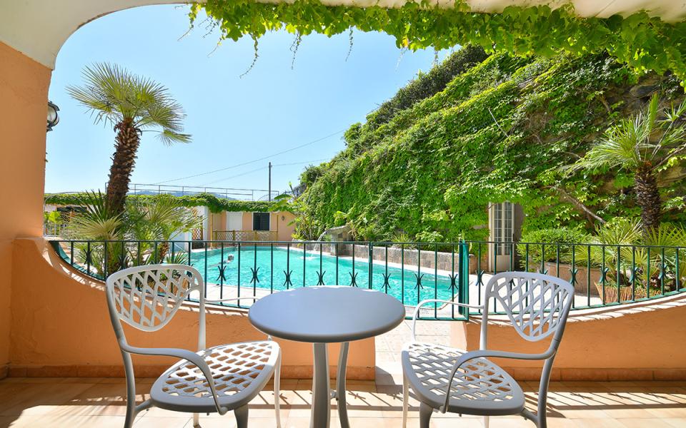 Terrazza con vista piscina, due sedie e tavolo, circondata da verde lussureggiante.