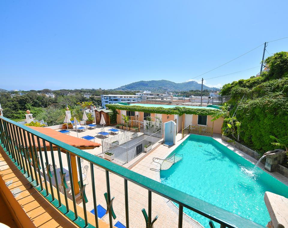 Piscina luminosa, ombrelloni bianchi, verde rigoglioso, cielo azzurro, colline serene all'orizzonte.