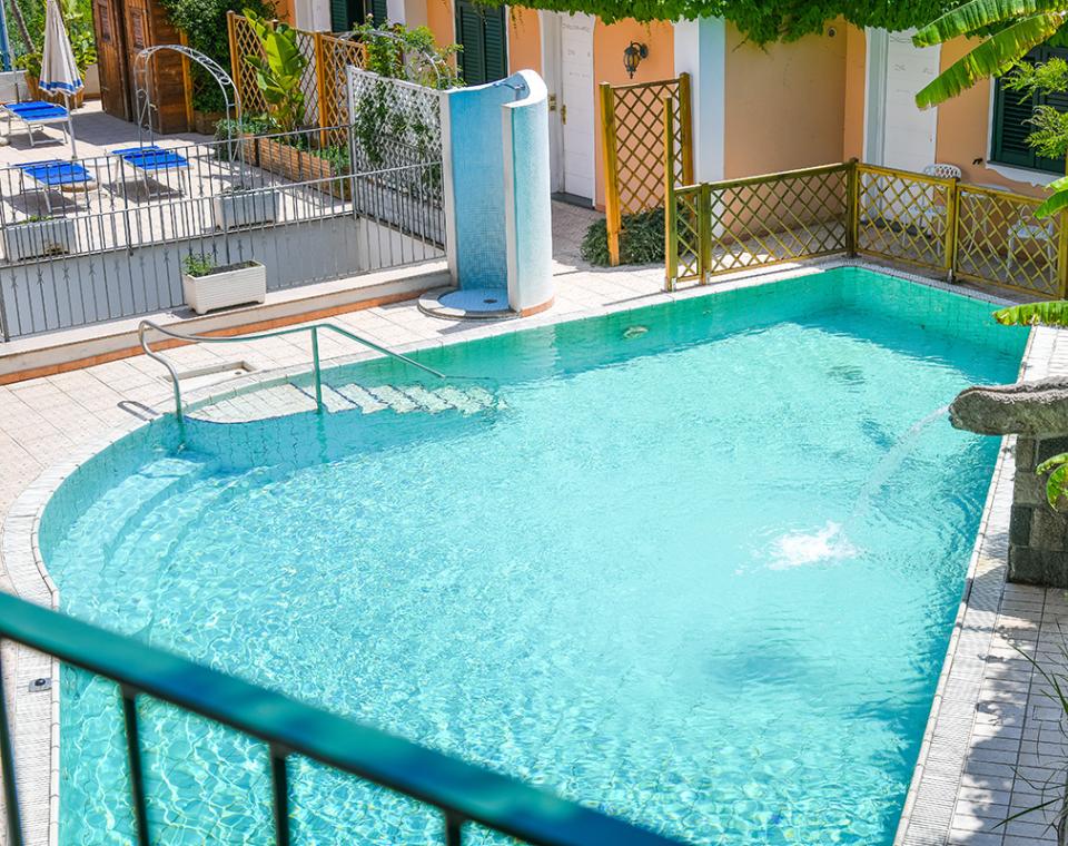 Piscina azzurra all'aperto, circondata da piante verdi e sedie a sdraio.
