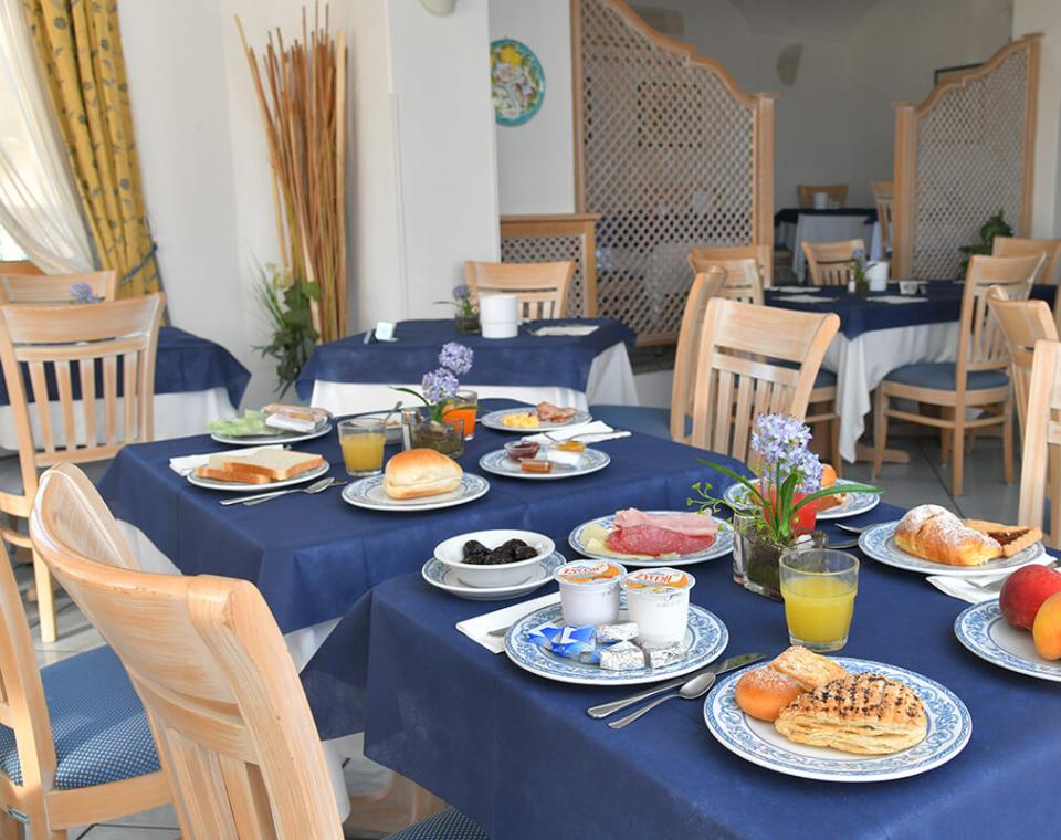 Colazione elegante in sala luminosa: frutta fresca, dolci, salumi e fiori colorati sui tavoli.