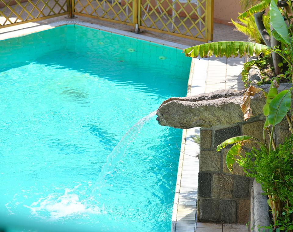 Piscina scintillante, getto d'acqua da roccia, circondata da piante lussureggianti, atmosfera tropicale.